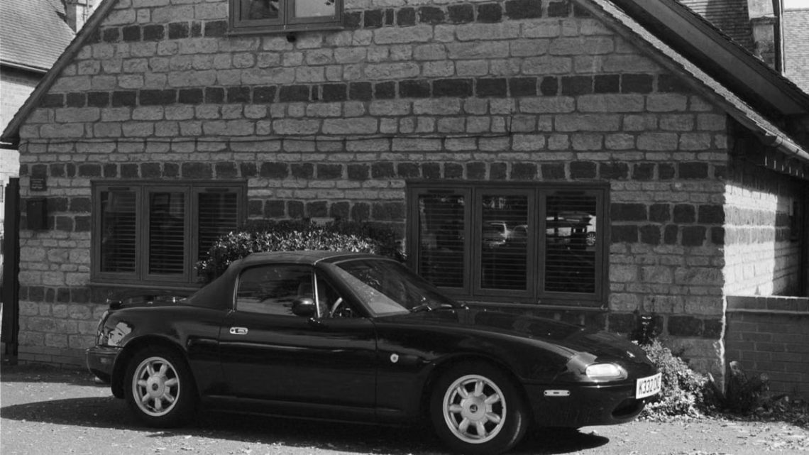 Black and white photo of a classic Mazda MX-5 Miata parked by a rustic stone house.