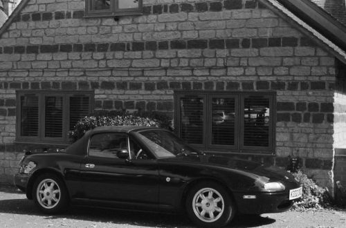 Black and white photo of a classic Mazda MX-5 Miata parked by a rustic stone house.