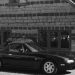Black and white photo of a classic Mazda MX-5 Miata parked by a rustic stone house.